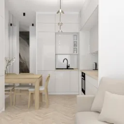 Jasne wnętrze z aneksem kuchennym, drewnianym stołem i krzesłami, oraz beżową sofą z poduszkami, oświetlone nowoczesną lampą wiszącą.