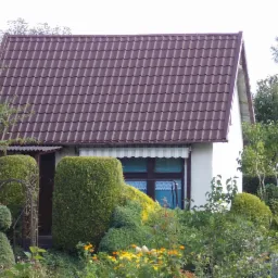 Dom jednorodzinny z brązowym dachem z blachodachówki w Elblągu, otoczony bujną zielenią i ozdobnymi krzewami.