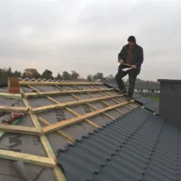 Dekarz na dachu z blachy trapezowej, mierzący odległość, widoczne łaty i kontrłaty, komin z czarnej cegły, pochmurne niebo w tle.