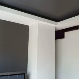 Nowoczesne wnętrze z szarą ścianą, białą kolumną i czarnym sufitem podwieszanym z białym obramowaniem, widoczne fragmenty drzwi i telewizora.