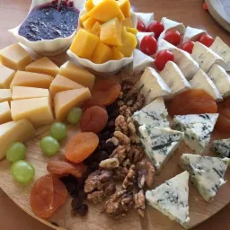 Deska serów z owocami i konfiturą na drewnianym talerzu: żółty ser, ser pleśniowy, morele, winogrona, orzechy włoskie, pomidorki cherry i miseczka z ciemną konfiturą.