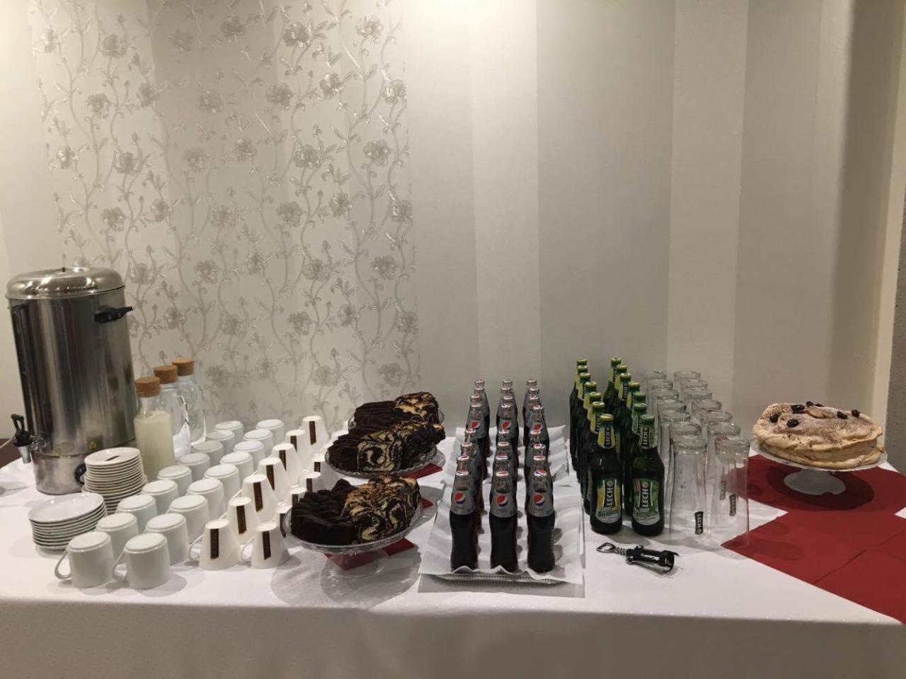 Stół z cateringiem: napoje w butelkach, kawa w termosie, ciasta marmurkowe i tort na tle ściany z kwiatowym wzorem.