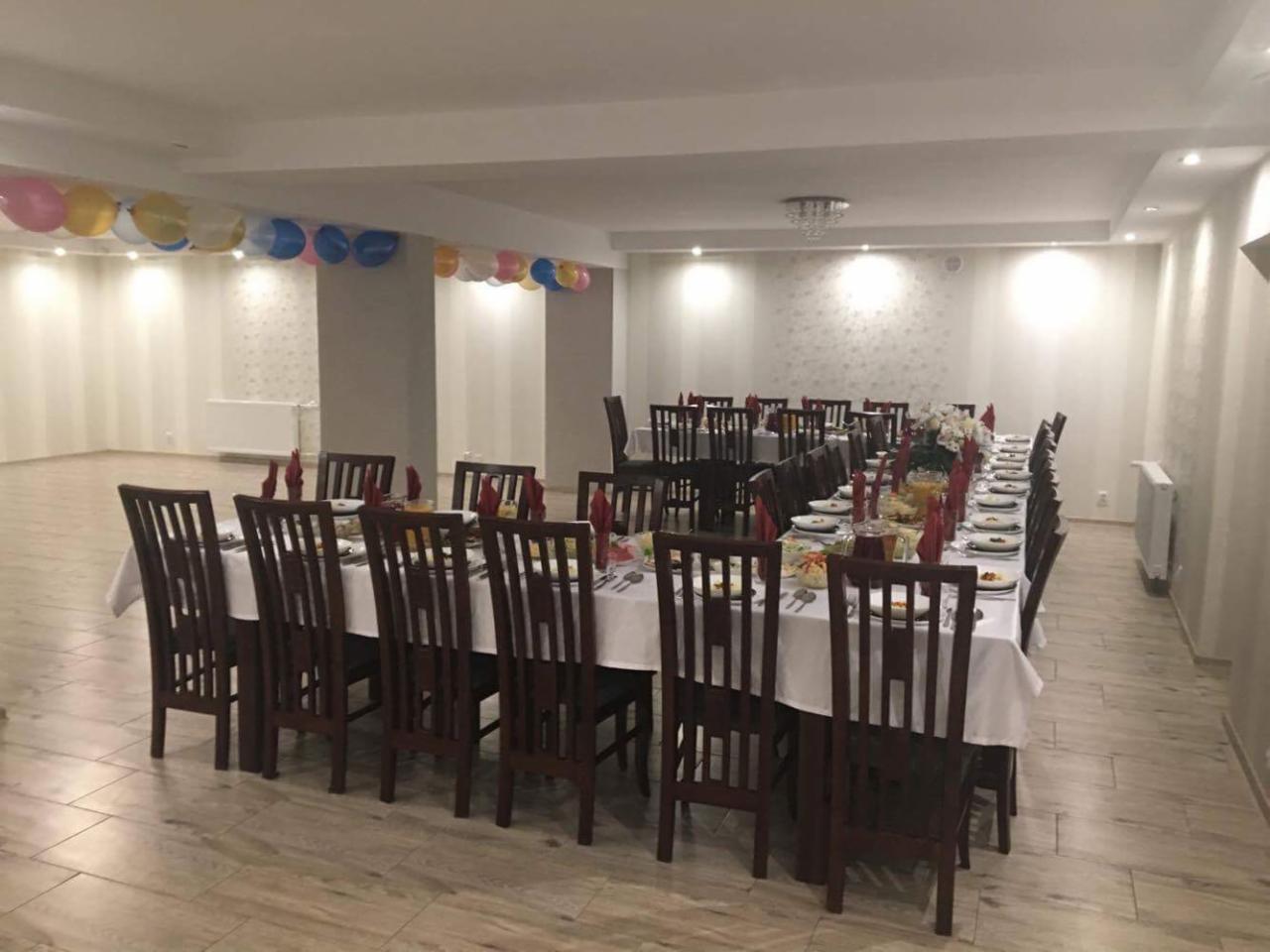 Uroczysta sala z długim stołem nakrytym białym obrusem, ciemne drewniane krzesła, zastawa stołowa, serwetki w kolorze czerwonym, dekoracja kwiatowa na środku stołu, balony pod sufitem.