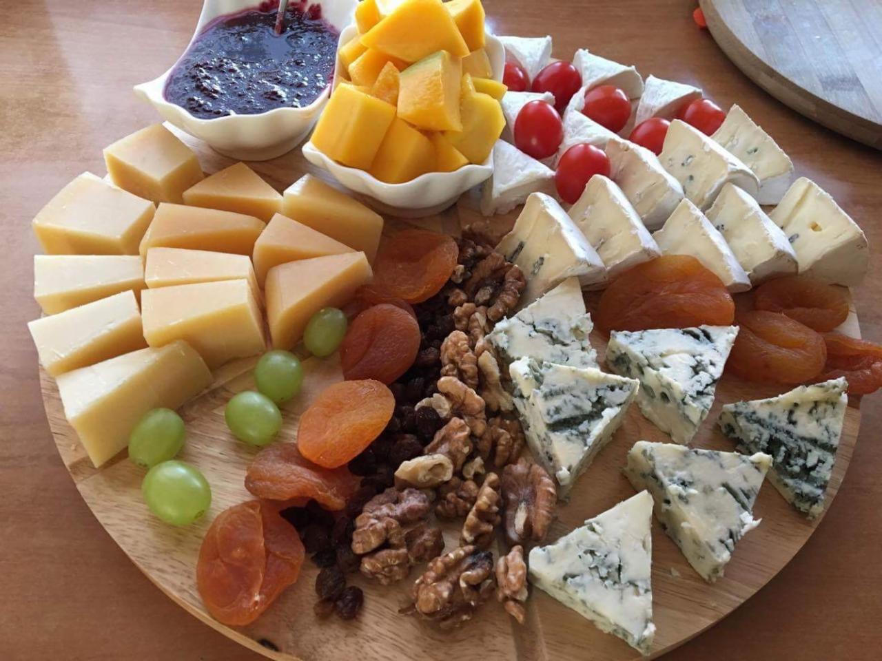 Deska serów z owocami i konfiturą na drewnianym talerzu: żółty ser, ser pleśniowy, morele, winogrona, orzechy włoskie, pomidorki cherry i miseczka z ciemną konfiturą.