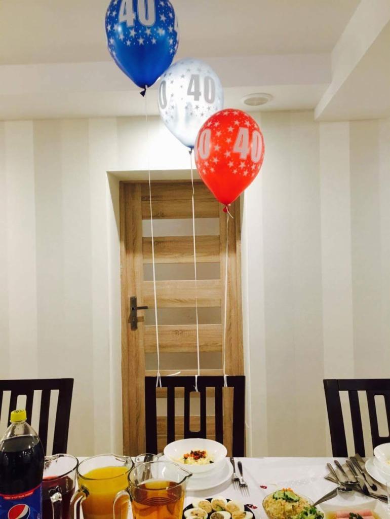 Uroczysty stół z napojami i przekąskami, nad nim unoszą się trzy balony w kolorach niebieskim, srebrnym i czerwonym z nadrukowaną liczbą 40, ustawione przed drewnianymi drzwiami.