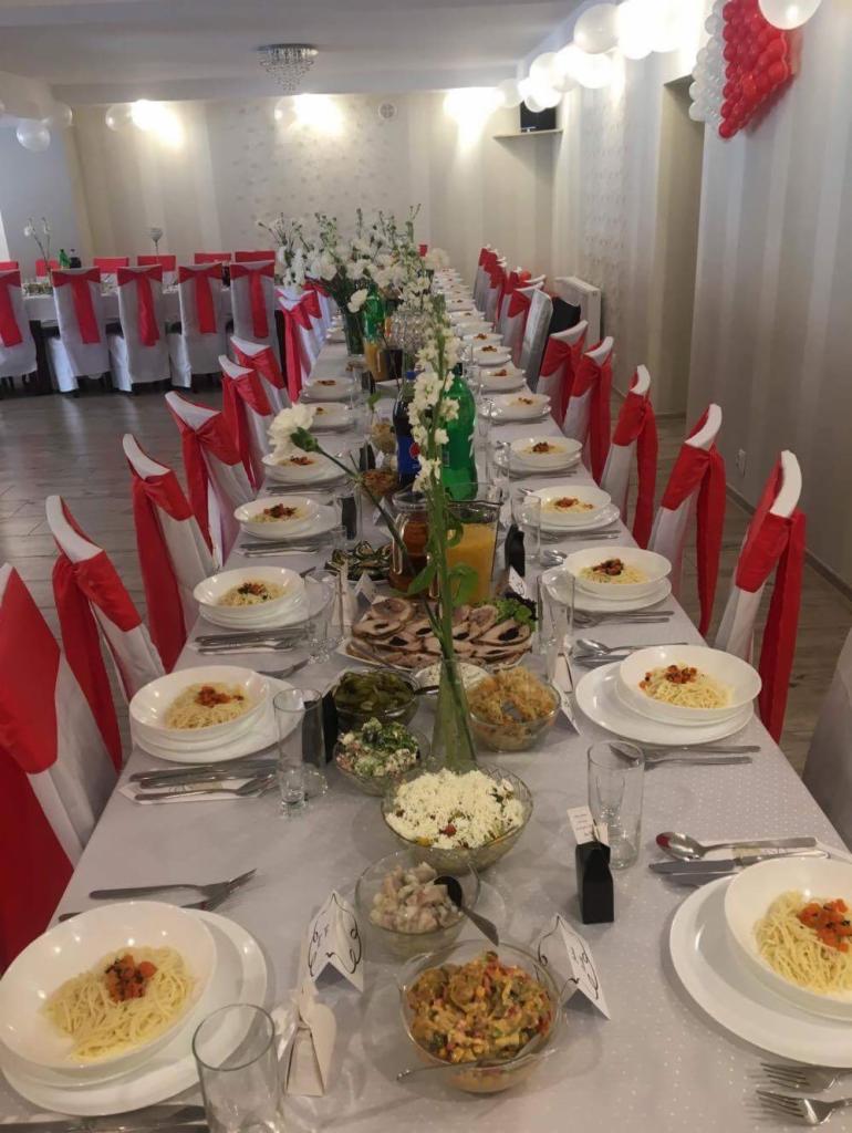 Uroczysty stół z białym obrusem, nakryty dla wielu gości, z potrawami w szklanych misach i talerzach, dekorowany białymi kwiatami w wazonach i balonami, krzesła z białymi pokrowcami i czerwonymi...