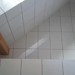 Wykończona łazienka na poddaszu w Koszalinie, z białymi, kwadratowymi płytkami ceramicznymi na ścianach, skośnym suficie i drewnianą belką konstrukcyjną.