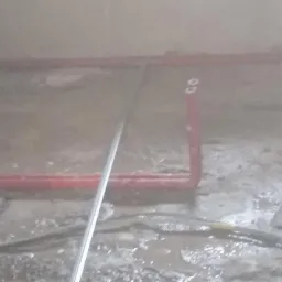 Surowy stan podłogi w trakcie remontu z widocznymi rurami grzewczymi w kolorze czerwonym, kablami i narzędziem do rozprowadzania zaprawy.