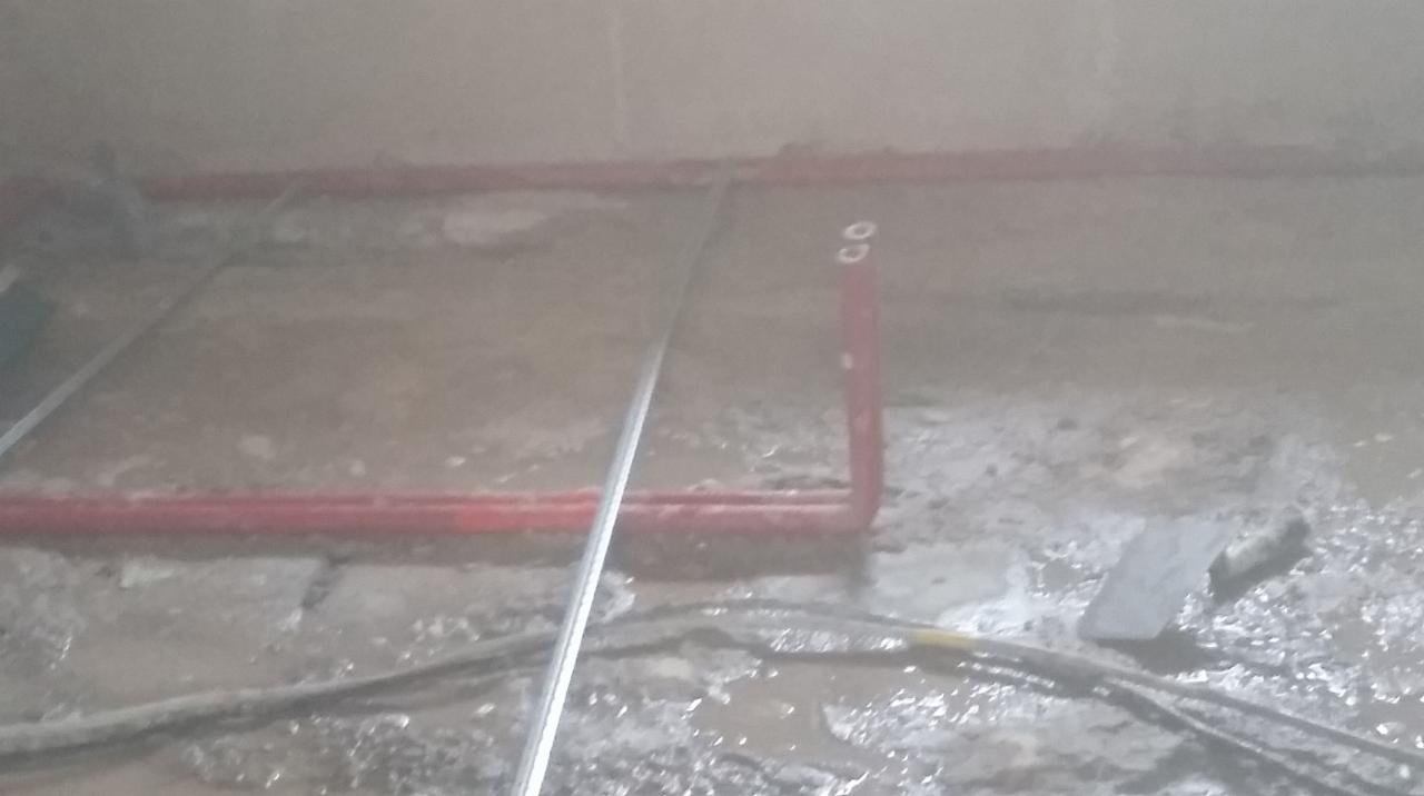 Surowy stan podłogi w trakcie remontu z widocznymi rurami grzewczymi w kolorze czerwonym, kablami i narzędziem do rozprowadzania zaprawy.