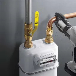 Instalacja gazomierza z użyciem zaciskarki do rur miedzianych, widoczny licznik gazu, rury miedziane i stalowe oraz żółty zawór odcinający.