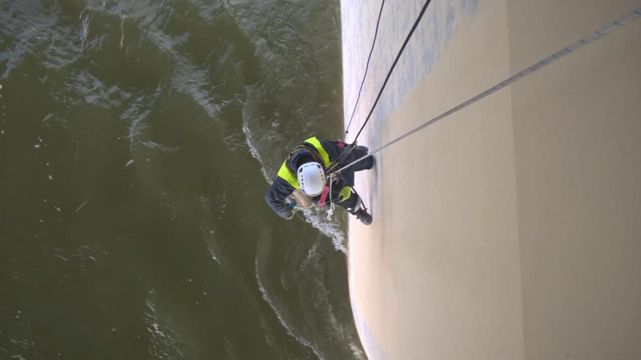 Pracownik w kasku i uprzęży, zawieszony na linach, wykonuje prace na wysokości na elewacji budynku nad wodą.