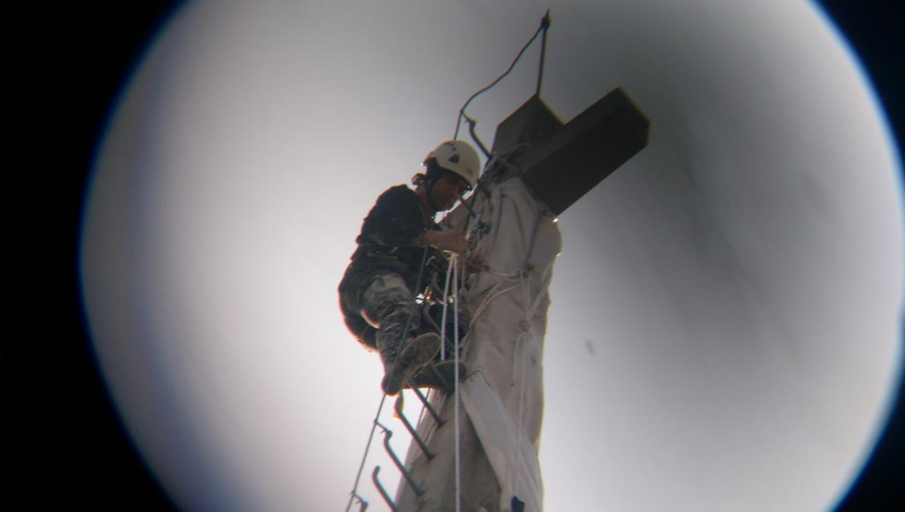 Pracownik w kasku na drabinie mocowanej do wysokiego, owiniętego materiałem krzyża, widok z dołu przez okrągły obiektyw, jasne tło.