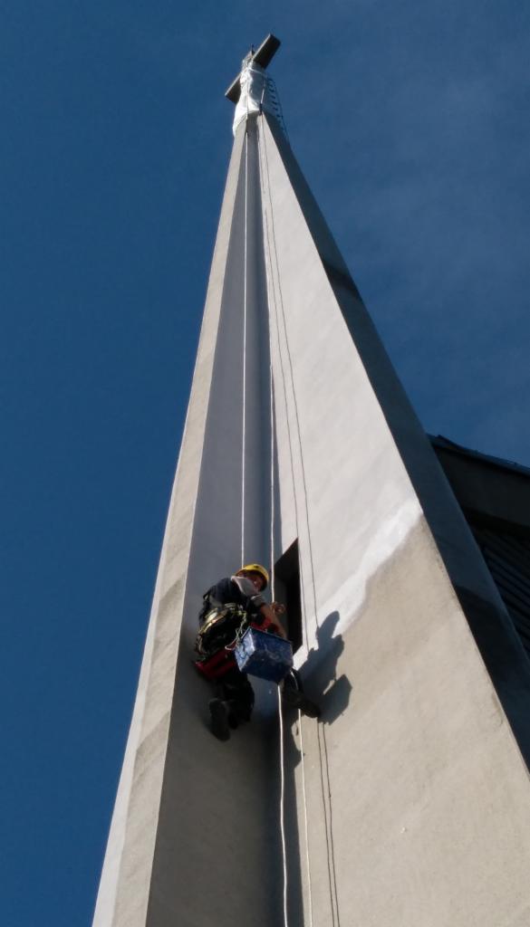 Pracownik w uprzęży, na wysokości, czyści elewację kościoła z krzyżem na szczycie, wisząc na linach obok małego okna.