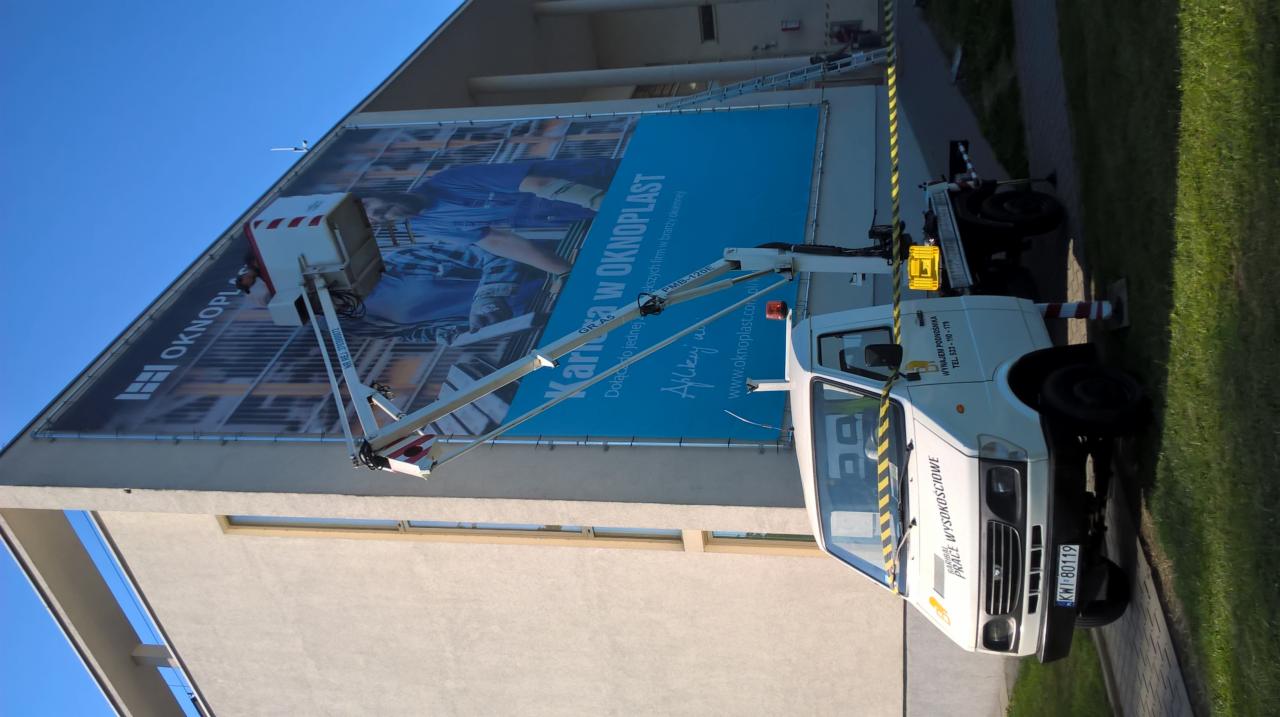 Biały samochód z podnośnikiem koszowym na tle budynku z dużym banerem reklamowym firmy Okoplast, widoczne detale konstrukcji podnośnika i oznaczenia na pojeździe.