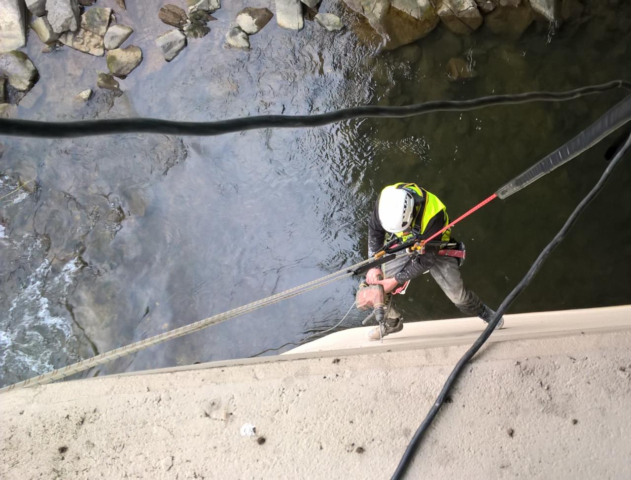 Pracownik w kasku i kamizelce odblaskowej, zabezpieczony linami, wierci w betonowej konstrukcji nad rzeką, widok z góry.