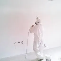 Pracownik w białym kombinezonie ochronnym maluje ścianę natryskowo, widoczne wystające kable elektryczne i wiadro z farbą.