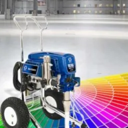 Agregat malarski natryskowy w kolorze niebieskim, marki Graco, z paletą barw w tle, na gładkiej posadzce w hali przemysłowej.