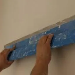 Nakładanie gładzi na ścianę przy użyciu szerokiej, niebieskiej szpachli, widoczne ślady materiału na narzędziu i dłoniach wykonawcy.