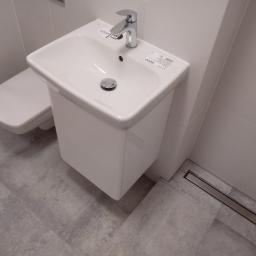 DSX sp. z.o.o - Biała umywalka z szafką, bateria i odpływ liniowy w nowoczesnej łazience z szarymi płytkami podłogowymi i wiszącą toaletą.