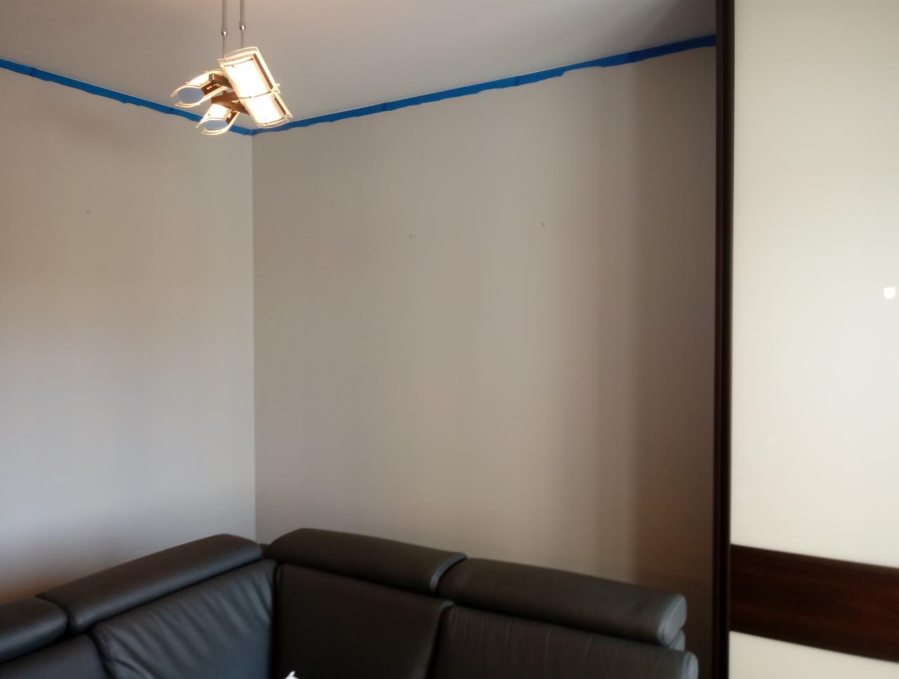 Przygotowanie do malowania ścian: ściany zabezpieczone niebieską taśmą malarską, widoczny fragment czarnej skórzanej sofy narożnej i przesuwna szafa z białymi frontami.