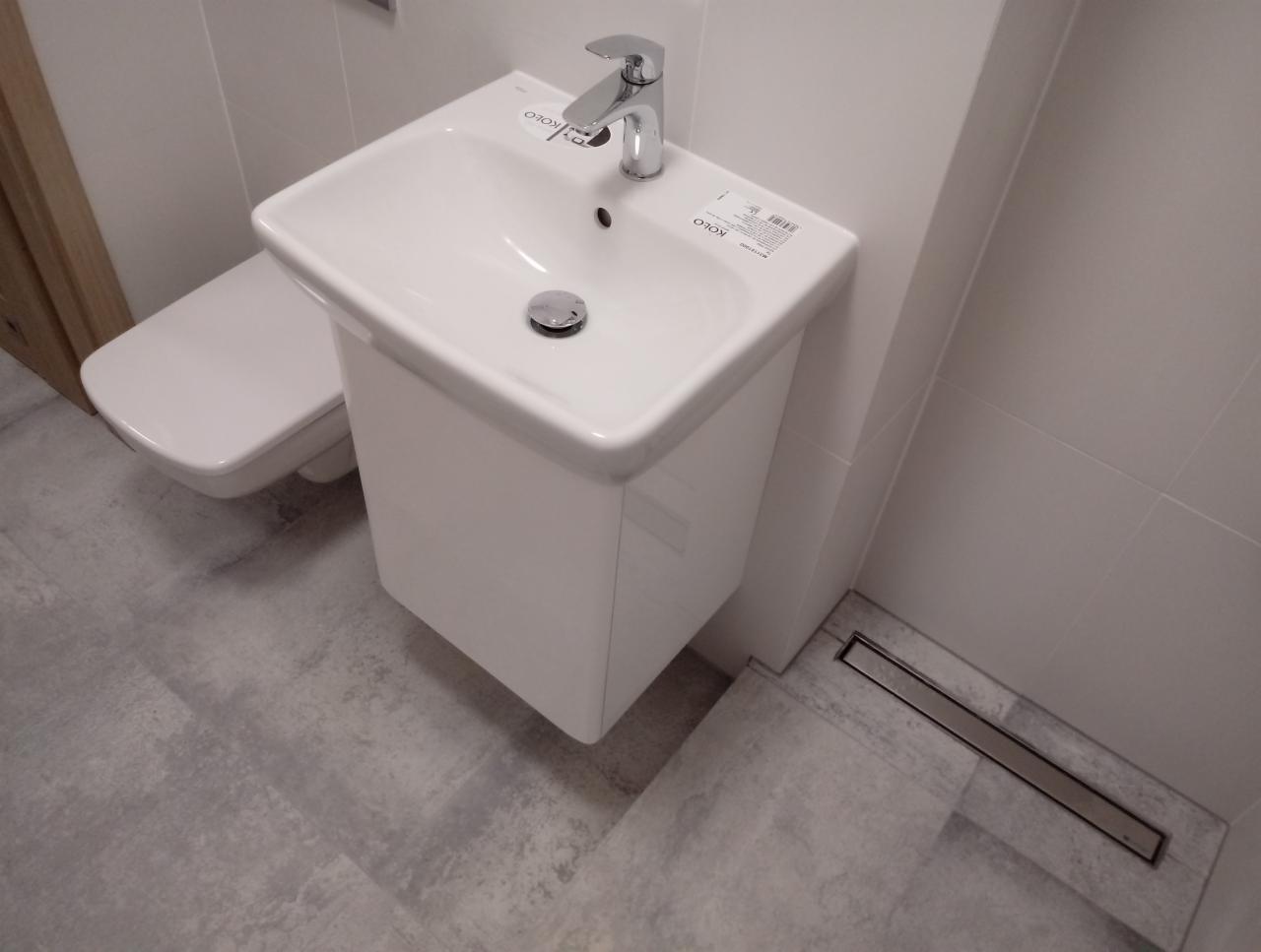 Biała umywalka z szafką, bateria i odpływ liniowy w nowoczesnej łazience z szarymi płytkami podłogowymi i wiszącą toaletą.