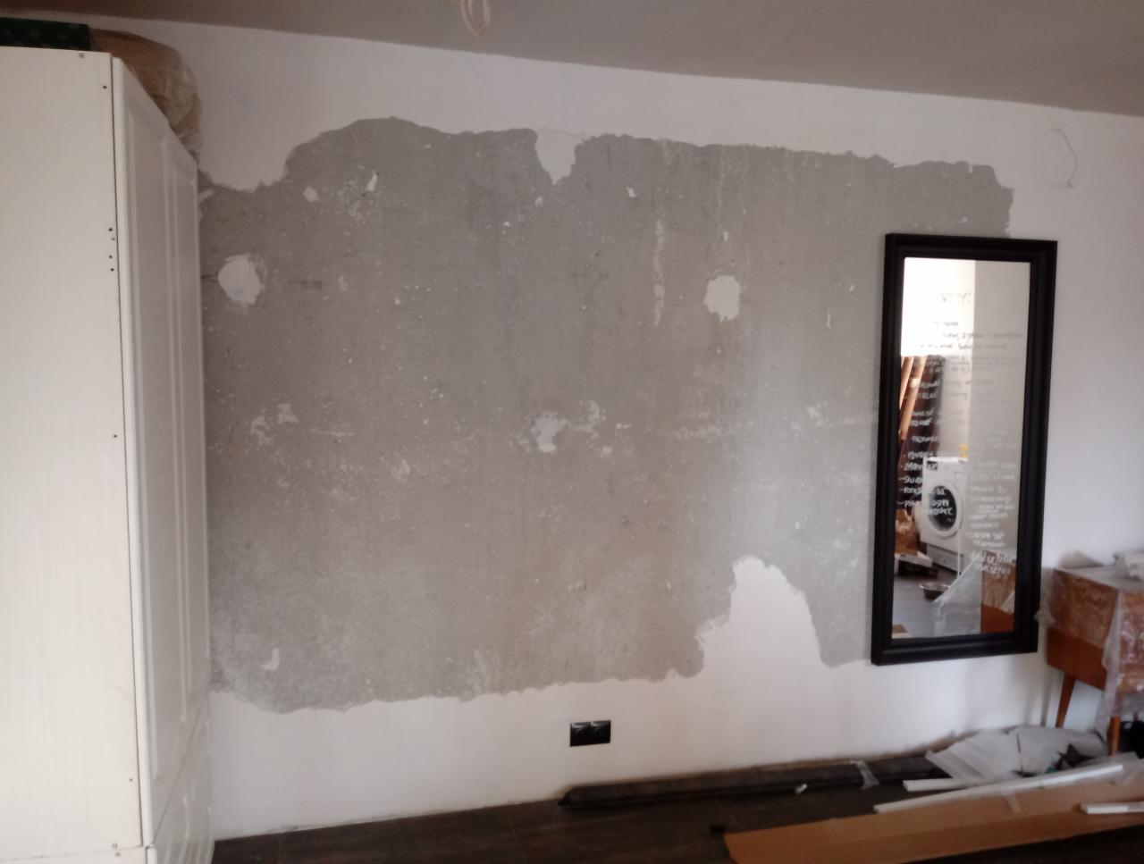 Przygotowanie ściany do malowania: usunięta stara warstwa farby, widoczne nierówności i ślady po otworach, biała szafka po lewej stronie i czarne lustro na ścianie po prawej.