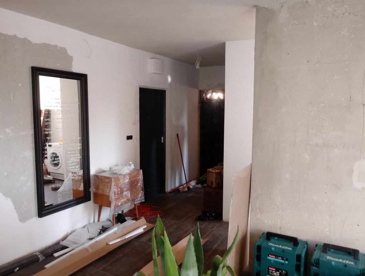 Wnętrze w trakcie remontu: surowe ściany, lustro z odbiciem pralki, zabezpieczona folią komoda, narzędzia i skrzynki Makita na podłodze.