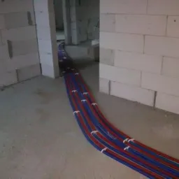 Rury grzewcze podłogowe, czerwono-niebieskie, ułożone na surowym betonie w pomieszczeniu z białymi ścianami z betonu komórkowego, zabezpieczone białymi uchwytami.