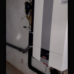 P.H.U. ANTA ZDZISŁAW PRZYGODZKI - Biały piec grzewczy marki Wolf z podłączonymi rurami i osprzętem hydraulicznym na tle surowej ściany w piwnicy.