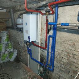 P.H.U. ANTA ZDZISŁAW PRZYGODZKI - Biały piec z wyświetlaczem, podłączony do instalacji z rur w izolacji czerwonej i niebieskiej, zamontowany na ceglanej ścianie w piwnicy, obok worki z pelletem.