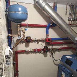 P.H.U. ANTA ZDZISŁAW PRZYGODZKI - Kotłownia z zainstalowanym kotłem na pellet, zbiornikiem wyrównawczym, pompami obiegowymi i izolowanymi rurami w kolorach czerwonym i niebieskim.