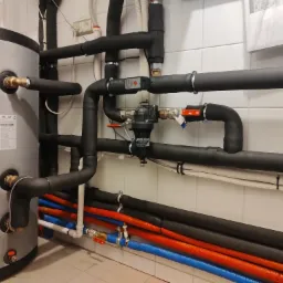 Szary zbiornik z podłączonymi rurami w izolacji, widoczne zawory, filtry i rury wodne w kolorze niebieskim, czerwonym i białym na tle białych, kwadratowych płytek.