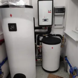 Dwa białe zbiorniki akumulacyjne ciepłej wody z czarnymi pokrywami, podłączone rurami do wiszącego kotła grzewczego marki Saunier Duval, zamontowane w białym pomieszczeniu technicznym.