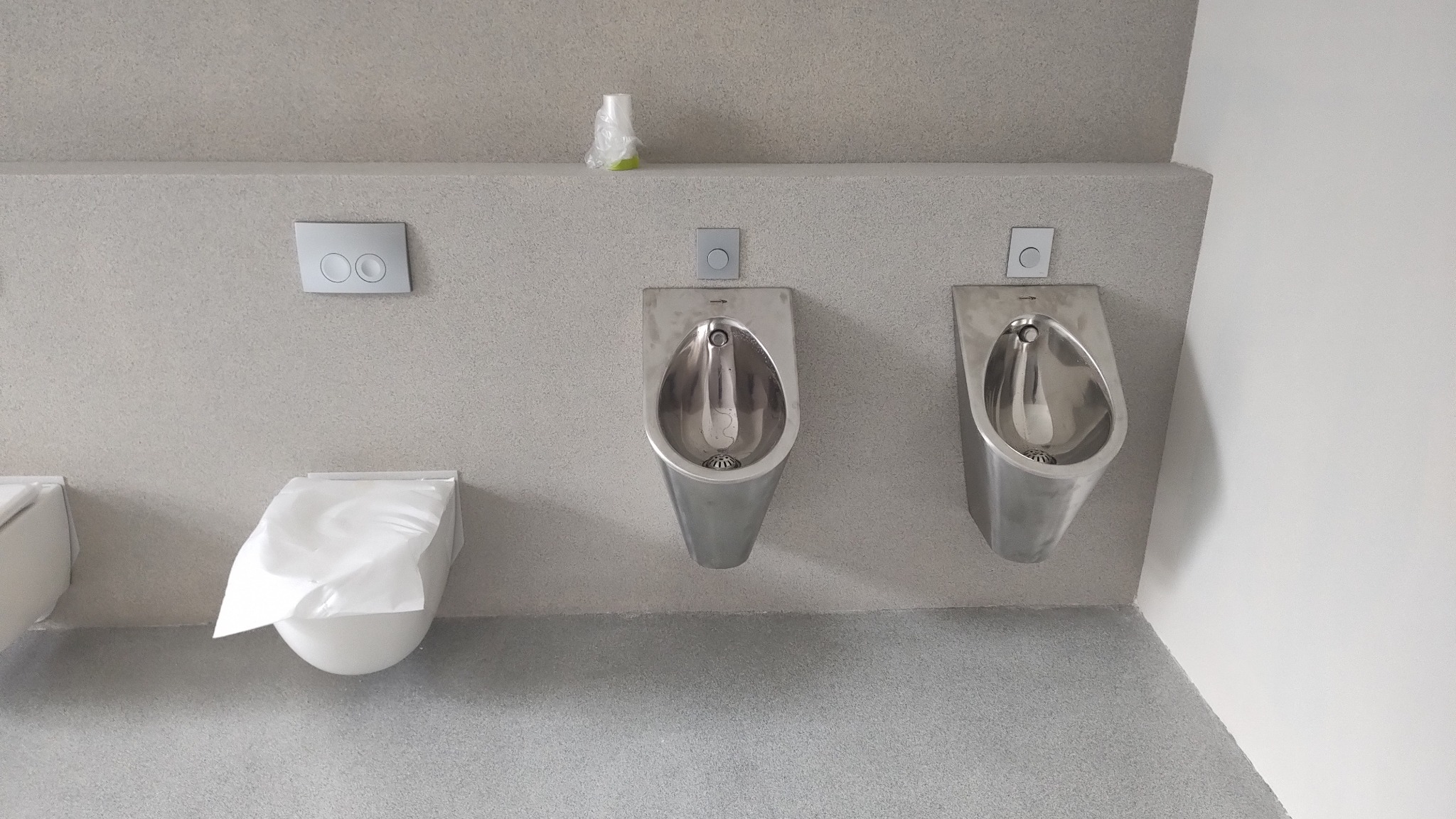 Dwa stalowe pisuary i toaleta pod przyciskiem spłukiwania w szarej łazience publicznej, zabezpieczone folią ochronną.