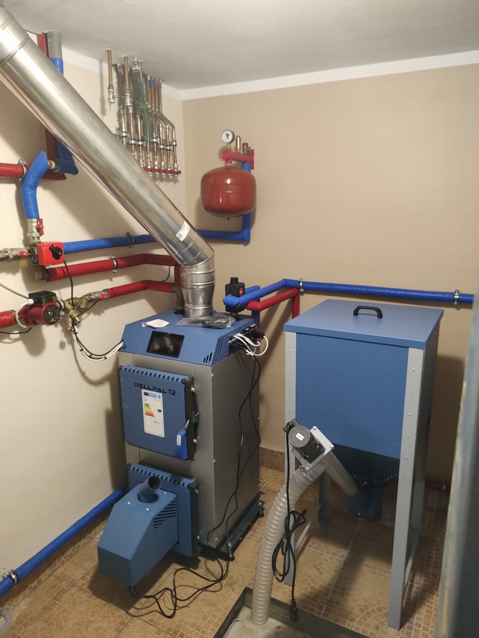 Kotłownia z kotłem na pellet PellPal 12, zasobnikiem, rurami instalacji CO i grzejnikami w tle, widoczne elementy instalacji hydraulicznej.