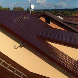 SPECMAL - Malowanie dachów na Podkarpaciu - Czysty, brązowy dach z blachy falistej na beżowym budynku w słoneczny dzień. Widoczne anteny i kominy na tle błękitnego nieba z chmurami.