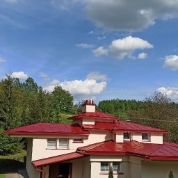 SPECMAL - Malowanie dachów na Podkarpaciu - Dom z czerwonym dachem na tle błękitnego nieba z chmurami i zieleni. Widok z góry ukazuje architekturę budynku i otaczający krajobraz.