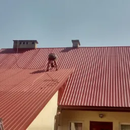 Pracownik w Krzywczy na dachu z blachy trapezowej w trakcie montażu zabezpieczeń, widoczne kominy i drabina oparta o ścianę budynku.