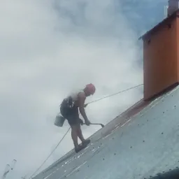 Pracownik w kasku, zabezpieczony linami, usuwa zabrudzenia z metalowego dachu przy użyciu narzędzia z długim uchwytem, widoczny komin i pochmurne niebo.