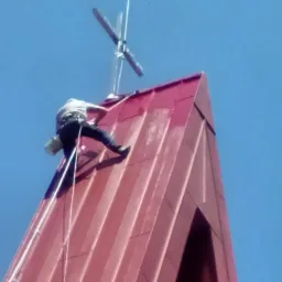 Pracownik na wysokościach, zabezpieczony linami, maluje czerwoną blachę na szczycie kościoła z krzyżem na tle błękitnego nieba.