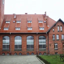 Ceglany budynek z wieżą zegarową i czerwoną dachówką, widoczne okna dachowe i duże okna łukowe z białymi ramami.