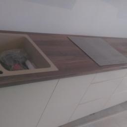 Wikbud - Fragment blatu kuchennego z drewnianym wzorem, z wbudowanym beżowym zlewem i płytą grzewczą, nad białymi szafkami z frezowanymi frontami.