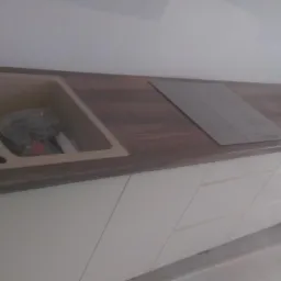 Fragment blatu kuchennego z drewnianym wzorem, z wbudowanym beżowym zlewem i płytą grzewczą, nad białymi szafkami z frezowanymi frontami.