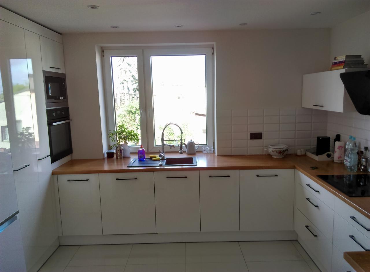 Jasna kuchnia z białymi, lakierowanymi frontami, drewnianym blatem i czarnymi uchwytami, widok na ogród przez okno nad zlewem.