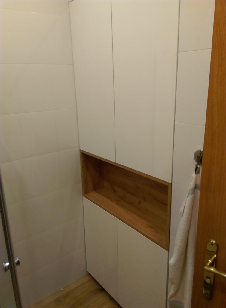 Biała szafa łazienkowa z drewnianą wnęką, zamontowana na ścianie z białych płytek, widoczny fragment drzwi z wieszakiem na ręcznik.