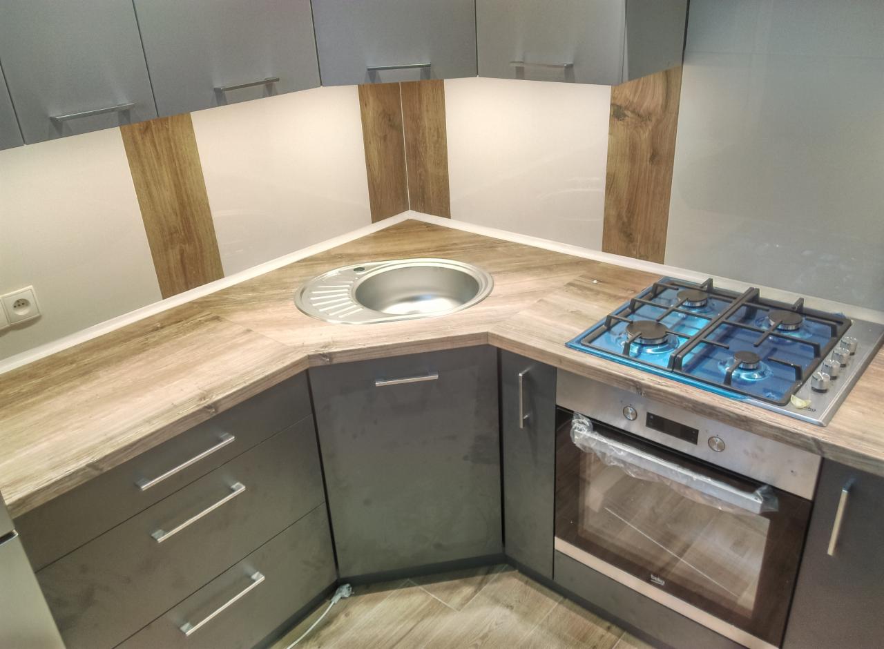 Szara, narożna zabudowa kuchenna z blatem imitującym drewno, okrągłym zlewem i płytą gazową z niebieską folią ochronną.