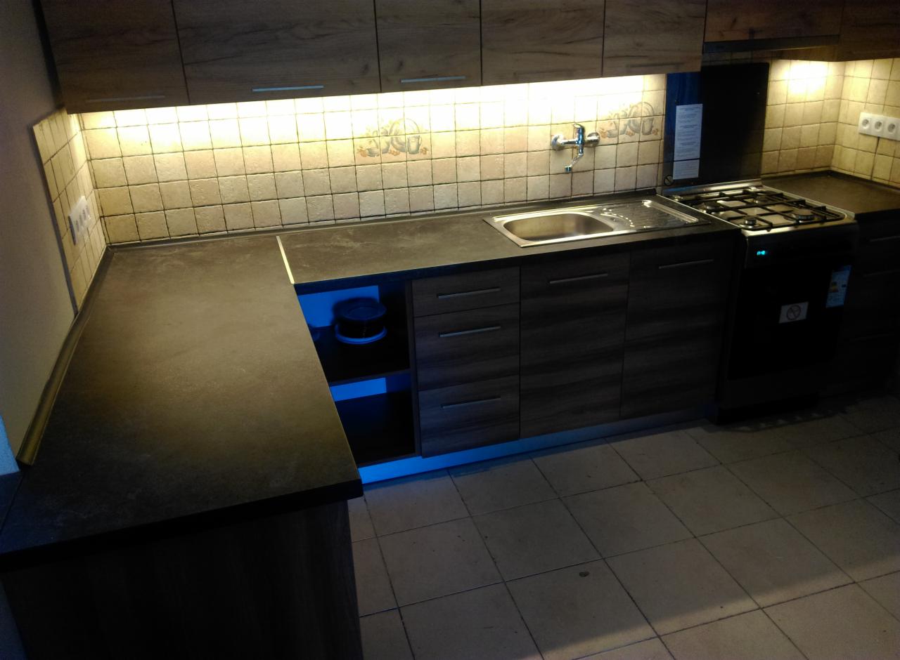 Ciemna, nowoczesna kuchnia z drewnianymi szafkami, podświetleniem LED i szarym blatem, widok na zlew i kuchenkę gazową.