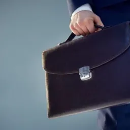 Mężczyzna w garniturze trzyma brązową skórzaną aktówkę z metalowym zapięciem na tle szarego gradientu.