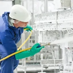 Pracownik w kombinezonie ochronnym i kasku myje pod ciśnieniem przemysłowe urządzenia pokryte pianą czyszczącą, widoczne przewody i dysza spryskująca, minimalizm i sterylność otoczenia.