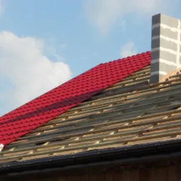 Montaż czerwonej blachodachówki na drewnianej konstrukcji dachu w Piotrków Trybunalski, widoczny komin w trakcie budowy i drabina oparta o rynnę.